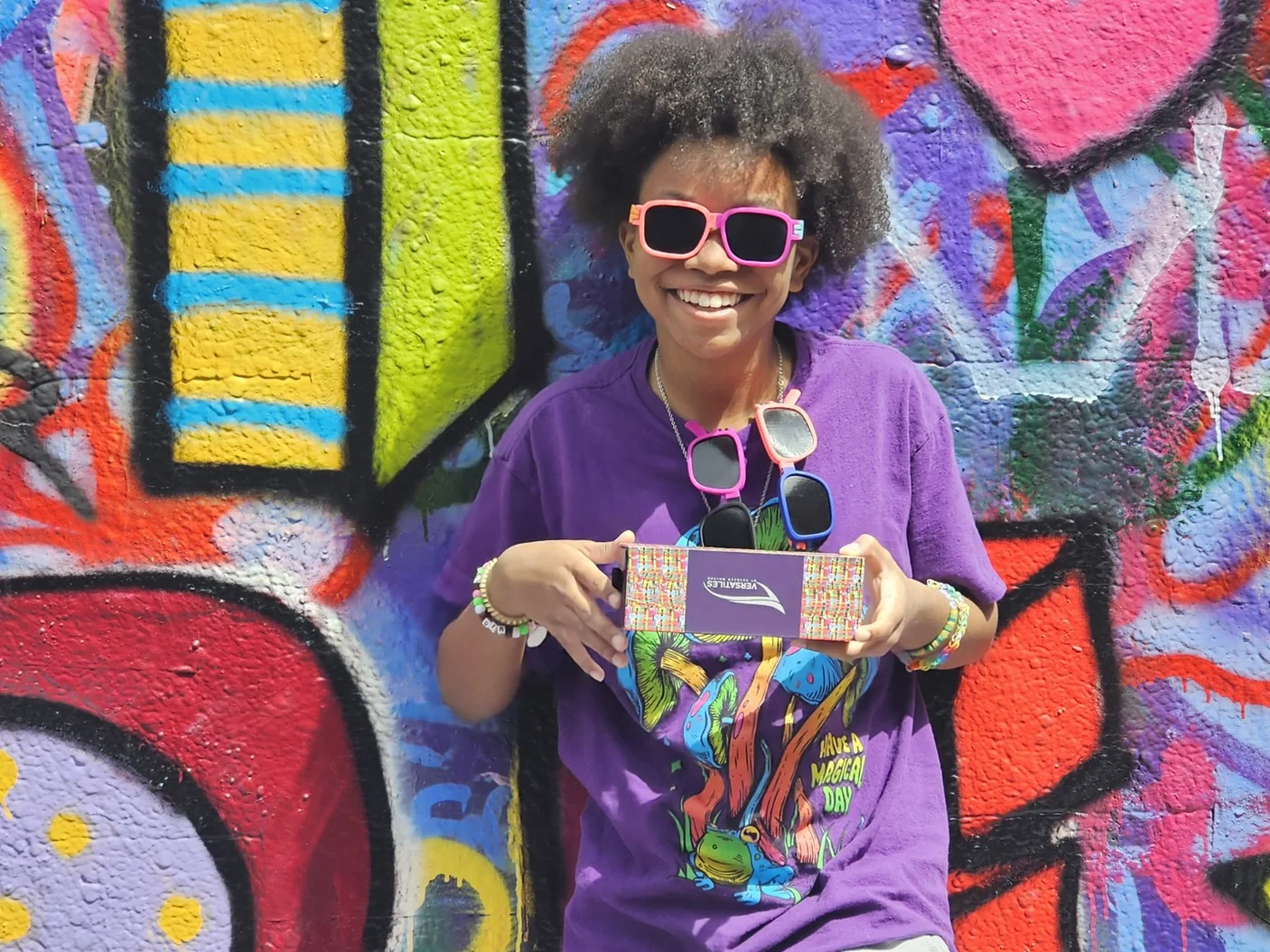 Child wearing pink and orange Versatiles sunglasses and holding the product box in front of a colorful mural